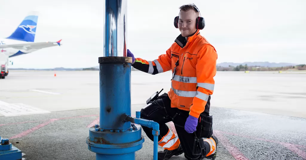 En lærling i oransje arbeidsklær står ved utstyr på en flyplass, med et fly i bakgrunnen.