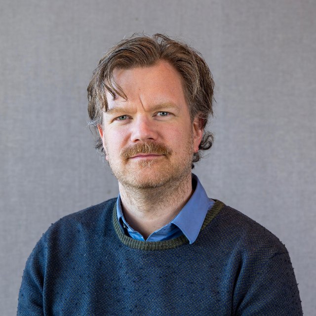Mann ser smilende inn i kamera, med blå genser og skjorte.