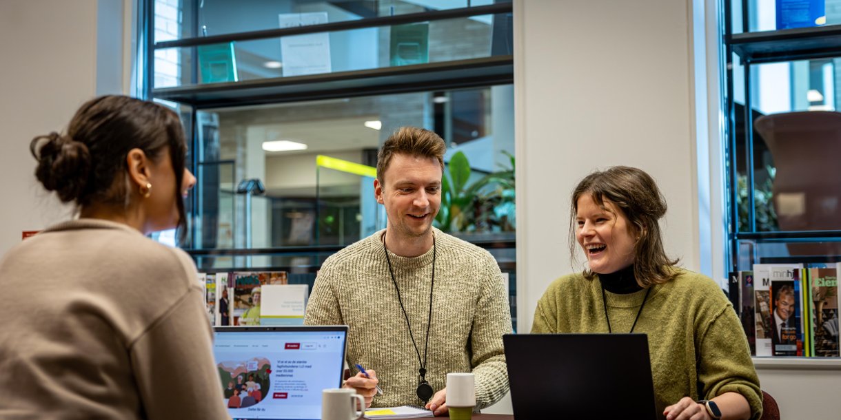 Tre personer sitter rundt et møtebord og smiler. På den ene PC-skjermen ser du NTLs nettsider.