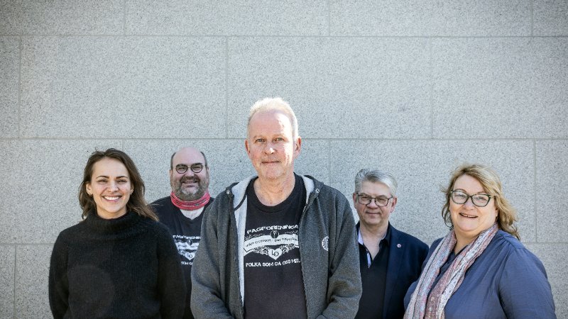 Fem personer står ved siden av hverandre foran en grå murvegg.