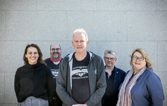 Fem personer står ved siden av hverandre foran en grå murvegg.