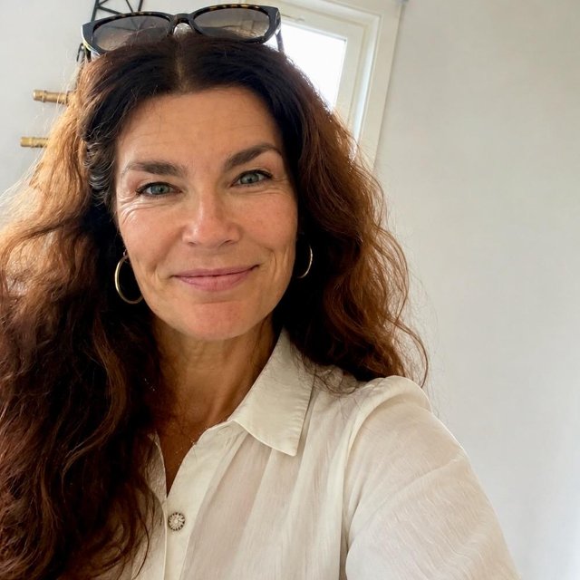 Kvinne ser smilende inn i kamera, hun står foran et vindu.