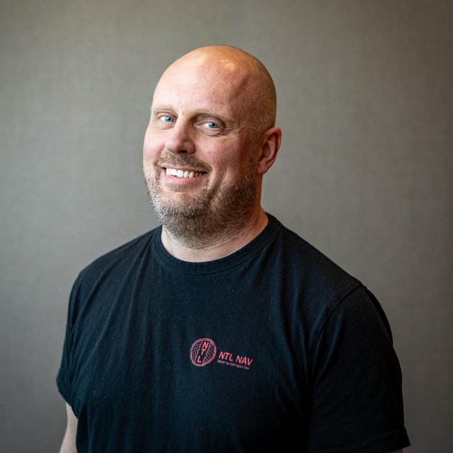 Mann ser smilende inn i kamera, han har på seg en svart t-skjorte og står foran en grå bakgrunn.