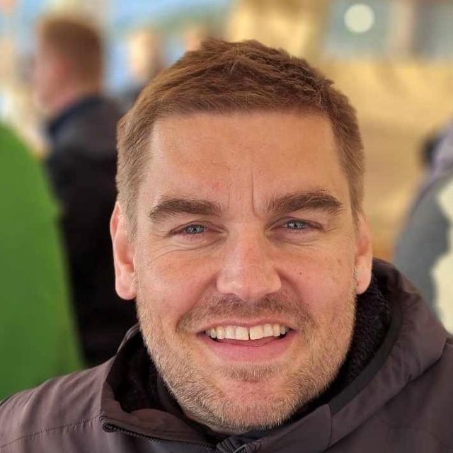 Mann med skjegg smiler, bak han er det andre mennesker som ikke er i fokus.